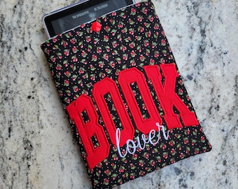May include: A black fabric sleeve with a red cherry and white polka dot pattern, featuring the red embroidered text BOOK and the white cursive text lover. A small red snap button secures the top, holding a digital e-reader device inside.