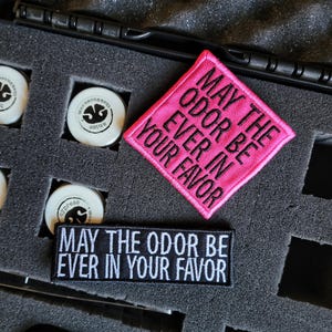 May include: Two embroidered patches with the text MAY THE ODOR BE EVER IN YOUR FAVOR. One patch is square and bright pink with black text, while the other is rectangular and black with white text. They are placed on dark gray foam padding.