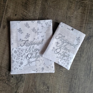 May include: Two rectangular fabric pouches in white with a grey floral and newspaper print pattern. Both items feature the embroidered text "Fictional Men Only" in a grey script font. The smaller pouch includes a single silver metal snap closure.