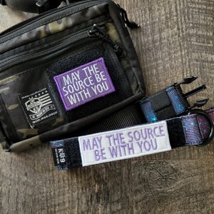 May include: A camouflage tactical bag with a purple rectangular patch reading MAY THE SOURCE BE WITH YOU and a matching galaxy-print dog collar featuring a white rectangular patch with the same text in purple lettering and a black plastic buckle.