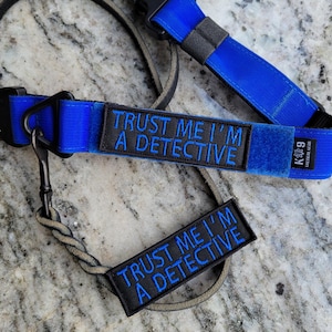 Puede incluir: Juego de correa y collar para perros azul con el texto "TRUST ME I'M A DETECTIVE" bordado en azul sobre un parche negro. El juego incluye un cierre de metal negro y un parche a juego.