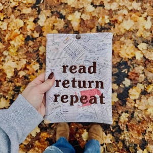 May include: A rectangular fabric pouch featuring a collage of vintage library checkout cards and receipts. The words "read return repeat" are embroidered in dark brown thread across the center. A single dark brown snap button secures the top.