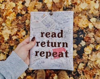 May include: A rectangular fabric pouch featuring a collage of vintage library checkout cards and receipts. The words "read return repeat" are embroidered in dark brown thread across the center. A single dark brown snap button secures the top.