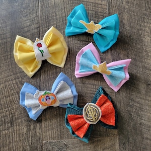 May include: Five colorful fabric hair bows with embroidered designs. The bows are in shades of yellow, blue, pink, and orange. Each bow features a different design, including a rose, a pumpkin carriage, and a magic lamp.