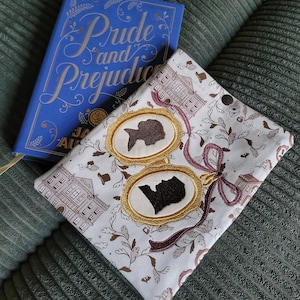 Könnte beinhalten: Ein blaues Hardcover-Buch mit dem Titel Pride and Prejudice neben einer weißen Stoff-Buchhülle mit einem Muster aus braunen Gebäuden und einem großen, gestickten goldenen Medaillon mit zwei dunkelbraunen Silhouetten, verziert mit einem weinroten Band.