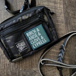 Puede incluir: Bolso bandolera negro y con estampado de camuflaje con un parche verde que dice "BIRCH & ANISE & CLOVE & CYPRESS". El bolso tiene una correa negra y un pequeño parche que dice "MADE IN USA". El bolso está sobre una superficie de madera.
