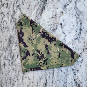 May include: A camouflage-patterned dog bandana in shades of green, tan, and black. The bandana features the name "LOKI" printed in black block letters on a sewn-on patch. The fabric has a pixelated digital design.