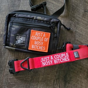 May include: A black camouflage tactical pouch with a rectangular orange patch featuring the white text JUST A COUPLE OF NOSY BITCHES. Attached is a red collar with a black buckle and a matching white embroidered patch with the same text.