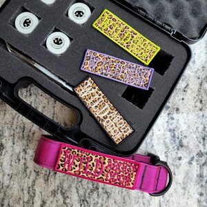 May include: A black carrying case containing three rectangular embroidered patches with leopard print and the text WILD ABOUT ODOR, a metal tool, and small white containers. A matching pink nylon collar with a leopard print patch is included.