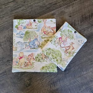 Könnte beinhalten: Zwei rechteckige Stoffbeutel mit einem sich wiederholenden Muster von Winnie Puuh-Figuren, darunter Puuh, Tigger und I-Aah in einer Waldumgebung mit grünen Bäumen, blauem Wasser und hellen Hintergründen auf einer weißen Stoffbasis.