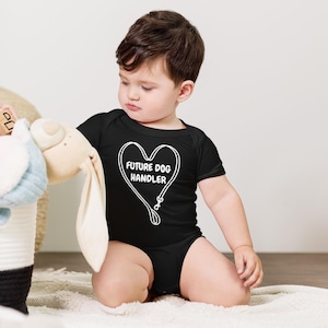 May include: A black baby bodysuit with a white heart-shaped graphic that includes a leash design and the text "FUTURE DOG HANDLER". The baby is kneeling on a white rug and holding a stuffed animal.