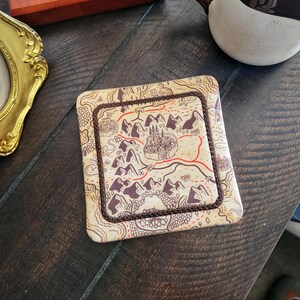 May include: Square coaster with a map design showing mountains, a castle, and roads in brown, red, and beige. It has a stitched brown border. The coaster is likely made of absorbent material, suitable for a cup or glass.