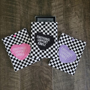 May include: Three rectangular protective sleeves featuring a black and white checkered pattern. Each sleeve has a heart-shaped patch with the text "EMOTIONAL SUPPORT KINDLE" in white. The patches are pink, black, and light purple respectively.