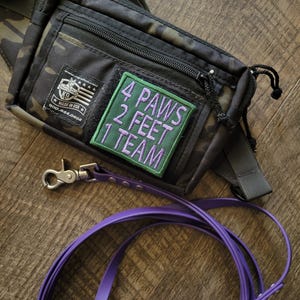 May include: A black camouflage waist bag with a rectangular patch featuring the purple text 4 PAWS 2 FEET 1 TEAM. The bag includes a small black label with white text and a K9 logo. A purple leash with a silver metal clasp is attached to the bag.