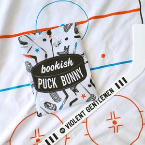 Puede incluir: Una funda para libros gris claro con un patrón de hockey que presenta patines, palos y cascos. La funda tiene un óvalo negro con las palabras "bookish PUCK BUNNY" en blanco. También se ve un palo de hockey blanco con el texto "VIOLENT GENTLEMEN".