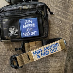May include: A black camouflage tactical pouch with a blue rectangular patch reading SNIFF AROUND AND FIND OUT, alongside a tan nylon collar featuring a matching rectangular patch with blue text reading SNIFF AROUND & FIND OUT and a black buckle.