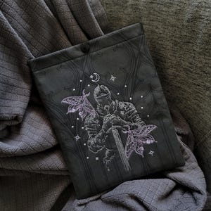 May include: A dark grey rectangular fabric sleeve featuring an embroidered design of a knight in armour holding a sword, flanked by two light purple moths. The background fabric has a subtle tonal gothic arch pattern and a single black button closure.