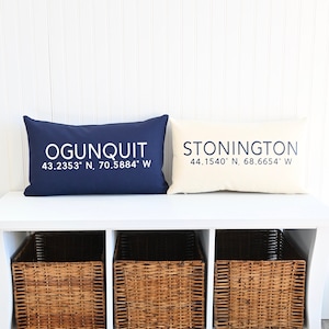 May include: A white bench with three wicker baskets underneath. Two decorative pillows are on top of the bench. The pillow on the left is navy blue with white text that reads "OGUNQUIT 43.2353° N, 70.5884° W". The pillow on the right is cream with navy blue text that reads "STONINGTON 44.1540° N, 68.6654° W".