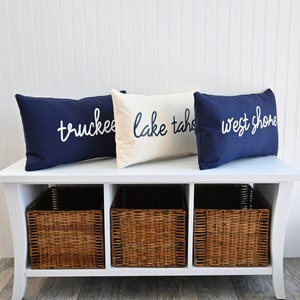 May include: White bench with three wicker baskets underneath. Three navy blue throw pillows with white text on them. The pillows read "truckee", "lake tahoe", and "west shore".
