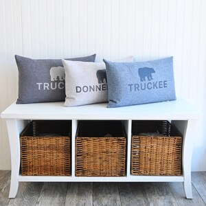 May include: White bench with three wicker baskets and three pillows. The pillows are gray, beige, and blue with the words "Truckee", "Donner", and "Truckee" printed on them.
