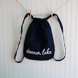 May include: A navy blue drawstring backpack with the words "donner lake" in white script. The bag is hanging from a clothesline with two wooden clothespins. The background is a white, vertical-slat wall.