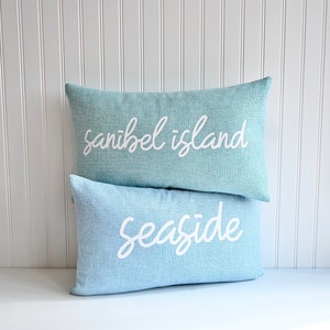 May include: Two light blue throw pillows with white text. The top pillow says "sanibel island" and the bottom pillow says "seaside".