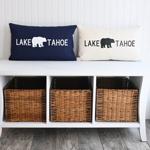 May include: A white bench with two pillows on top. The pillows are navy blue and white with the text "LAKE TAHOE" and a bear silhouette. The bench has three wicker baskets underneath.
