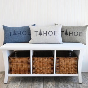 May include: A white bench with three woven baskets underneath. Three pillows are on top of the bench, each with the text "TAHOE" and a pine tree graphic. The pillows are in blue, gray, and light gray.