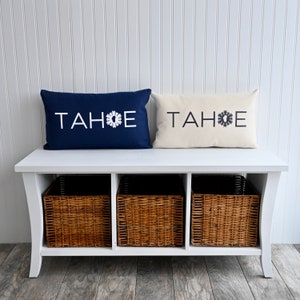 May include: White bench with three wicker baskets underneath. Two decorative pillows are on top of the bench, one navy blue with white text "TAHOE" and a snowflake, the other cream with the same text and snowflake.