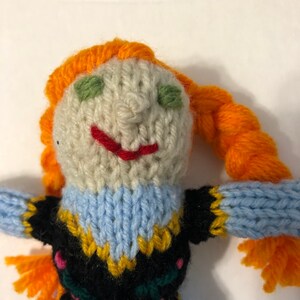 May include: A knitted doll with orange hair, a blue and black dress, and a white face with green eyes and a red smile.