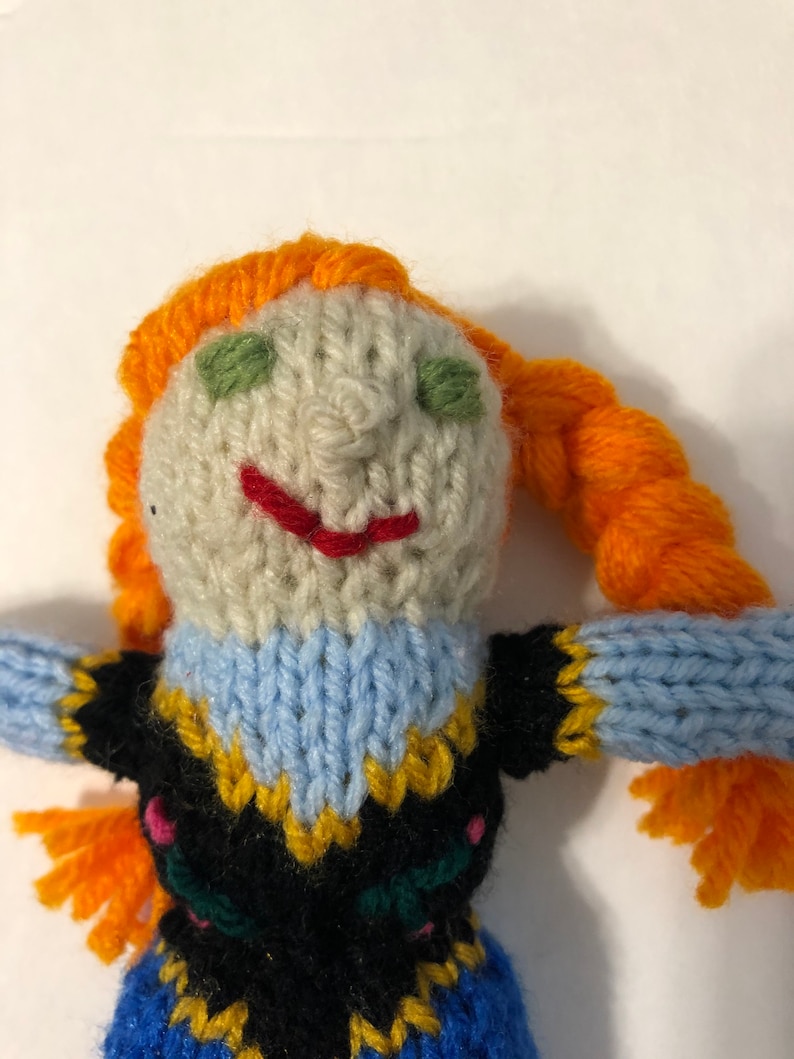 May include: A knitted doll with orange hair, a blue and black dress, and a white face with green eyes and a red smile.