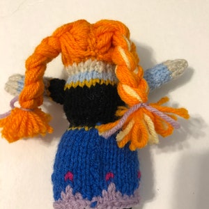 May include: A knitted doll with orange hair, a black bodice, a blue skirt, and purple trim. The doll has a braided hairstyle with orange and yellow yarn.