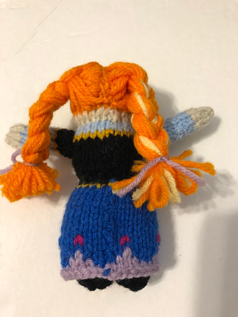 May include: A knitted doll with orange hair, a black bodice, a blue skirt, and purple trim. The doll has a braided hairstyle with orange and yellow yarn.