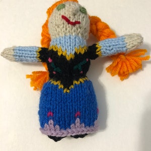 May include: A knitted doll with orange hair, a blue dress, and a black bodice with yellow and green embroidery. The doll has a white face with green eyes and a red smile.
