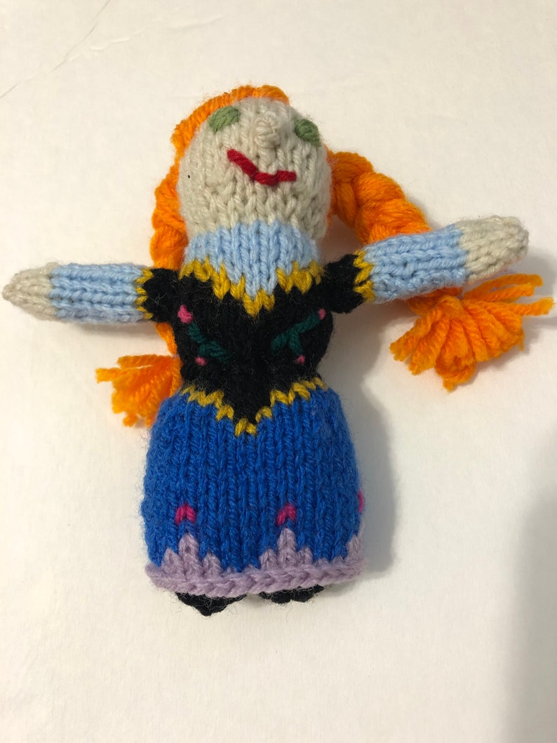 May include: A knitted doll with orange hair, a blue dress, and a black bodice with yellow and green embroidery. The doll has a white face with green eyes and a red smile.