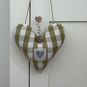 May include: A heart-shaped fabric decoration with a green and white gingham pattern, featuring a small gray heart applique. It hangs from a brown cord and has wooden button accents and a metal spring.