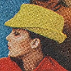 Op de afbeelding: Een close-up van een persoon die een gele hoed draagt. De hoed is een fedora-stijl met een brede rand. De persoon draagt een rood jack.