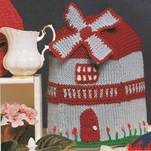 May include: A knitted teapot cozy shaped like a windmill with a red roof, blue walls, and a white window. The cozy has a red door and green grass with pink, yellow, and blue tulips.