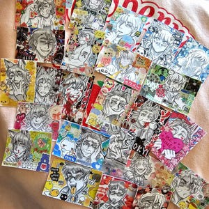May include: A collection of small, square art prints featuring anime-style character illustrations in black and white. Some prints include colorful stickers and text like "LOVE," "CANDY," and "Have a good day." The prints are arranged on a pink surface.