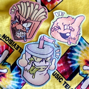 May include: Three cartoon stickers featuring food-themed characters. One sticker depicts a carton of fries with an angry face, another a pink brain with a goofy expression, and the last a soda cup with a straw and a face. The stickers are on a yellow tie-dye background.