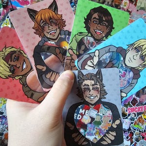 Może przedstawiać: Dłoń trzymająca pięć kart z grafikami postaci. Każda karta przedstawia inną postać w stylu anime z wilczymi uszami. Karty mają tła z r&oacute;żowych serc, zieleni i błękitu. Centralna karta ma serce z napisem "ME ED" i kolorowymi naklejkami z misiami.