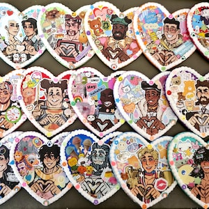 May include: A collection of heart-shaped coasters with colorful illustrations of various characters. Each coaster features a unique design with a white border and is decorated with stickers and drawings. The coasters are arranged in rows, showcasing a variety of artistic styles.