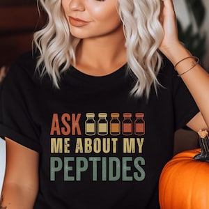 以下が含まれることがあります： 胸の中央にレトロなフォントで「ASK ME ABOUT MY PEPTIDES」というテキストがプリントされた黒の半袖Tシャツ。テキストの上には、黄色、オレンジ、赤の様々な色合いの小瓶のグラフィックイラストが5つ並んでいます。