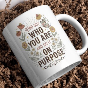 May include: A white ceramic mug featuring a floral and butterfly design with the text "FIND OUT WHO YOU ARE AND DO IT ON PURPOSE" in brown lettering. The illustration includes orange, yellow, and green botanical elements surrounding the quote.