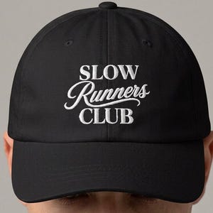 May include: A black baseball cap featuring white embroidered text on the front that reads SLOW Runners CLUB. The cap has a structured crown, a curved brim, and a button at the top center. The design is centered on the front panel of the hat.