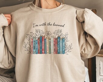 Può includere: Felpa beige con la scritta "I'm with the banned" sopra una fila di libri colorati. I libri sono decorati con illustrazioni floreali e di foglie. La felpa è di colore neutro.