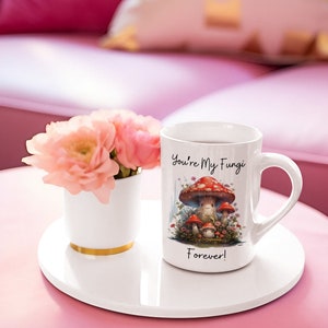 May include: White ceramic mug with a mushroom illustration and the text "You're My Fungi Forever!" The mug is on a white coaster and a pink surface.
