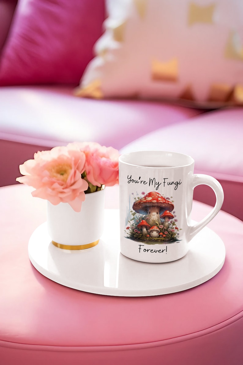 May include: White ceramic mug with a mushroom illustration and the text "You're My Fungi Forever!" The mug is on a white coaster and a pink surface.