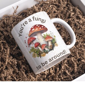 May include: White ceramic mug with a brown handle. The mug has a colorful illustration of mushrooms and the text "You're a fungi to be around!"