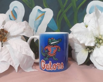 Puede incluir: Una taza de cerámica azul con un cocodrilo de dibujos animados que lleva una camisa roja y azul con la letra "F" en ella. La taza dice "Gators" en letras blancas.